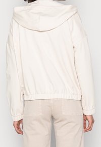 Světle béžová bunda s kapucí a volným střihem, elastickým páskem a žebrovanými manžetami. Hladká textura s minimem švů a bez viditelného brandingu.