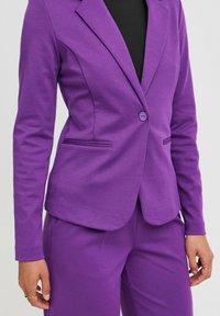 Blazer sur mesure violet avec une fermeture à un bouton, deux poches avant et un tissu texturé subtil. Design ajusté avec des manches longues.