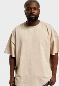 Beige, oversized, rövid ujjú póló puha anyagból, kerek nyakkivágással és finom tonális logórészlettel a gallér közelében.