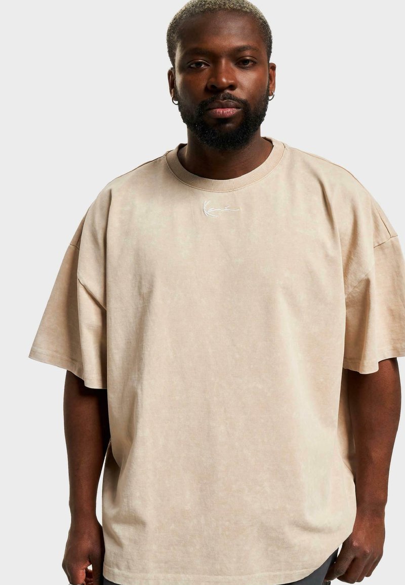 Beige, oversized, rövid ujjú póló puha anyagból, kerek nyakkivágással és finom tonális logórészlettel a gallér közelében.