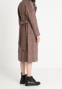 Cappotto lungo a quadri in tonalità di beige, rosso e nero. Presenta una vita con cintura e due tasche laterali. Stivali neri in pelle alla caviglia completano l'outfit.
