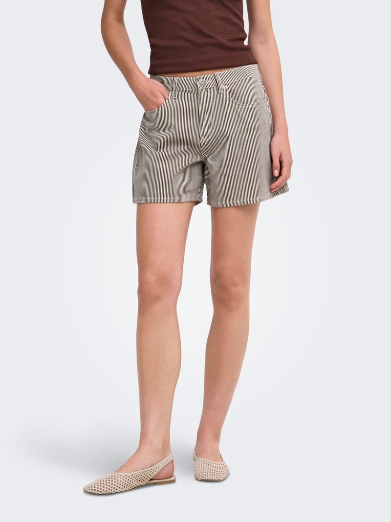 Femme portant un short beige et blanc à rayures verticales, un haut marron sans manches et des chaussures plates tissées beige, debout avec une main dans la poche.