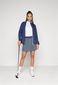 Žena v námořnickém saku, bílé zip-up košili, vzorované sukni, bílých ponožkách a teniskách, držící golfovou hůl, stojící na jednobarevném šedém pozadí.