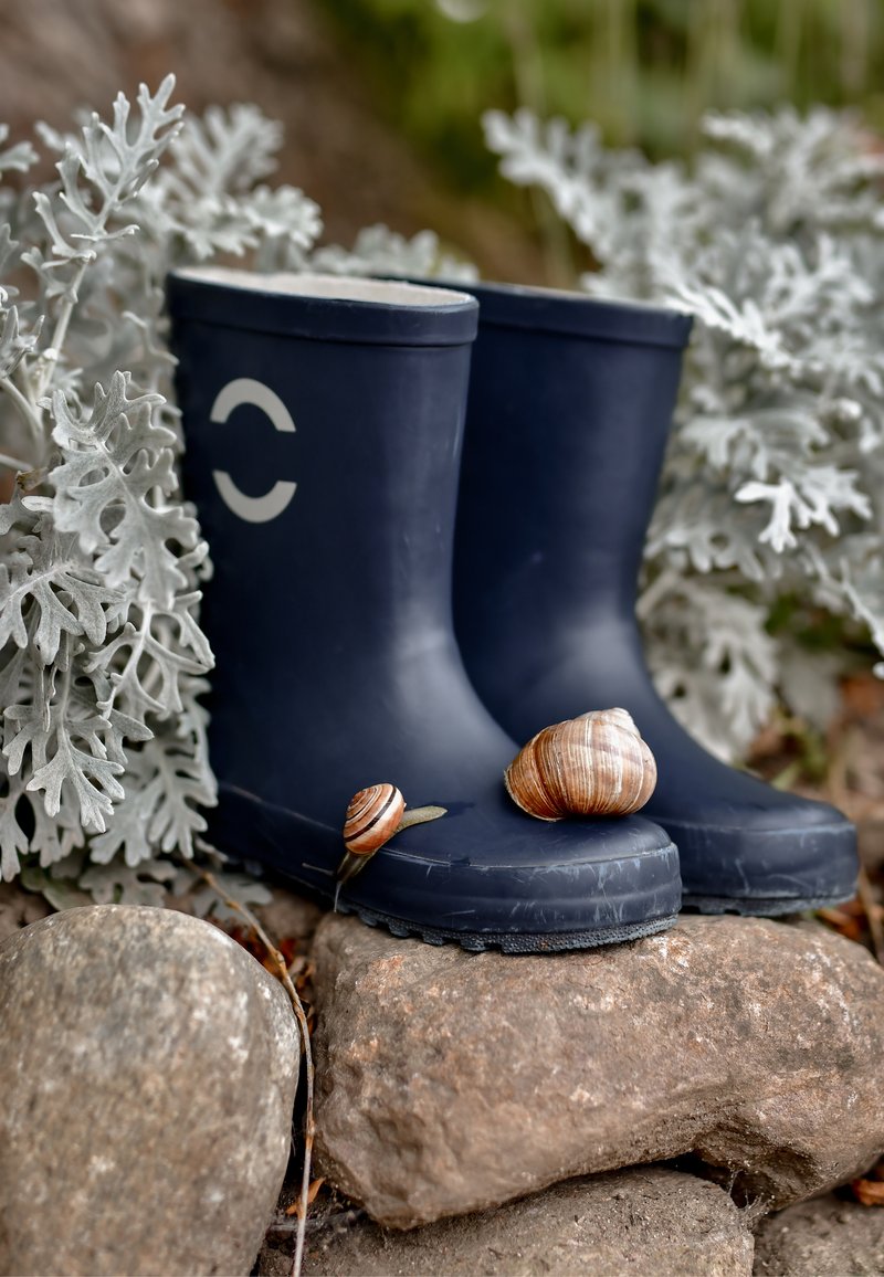 Niebieskie gumowe kalosze o zaokrąglonym kształcie z białym logo, ustawione na kamieniach, obok dwa ślimaki, otoczone zielonym listowiem.