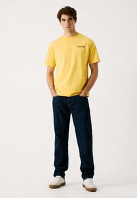 Gul bomulds T-shirt med korte ærmer og rund hals, der har et lille logo på brystet. Sammen med mørkeblå jeans og hvide sneakers.
