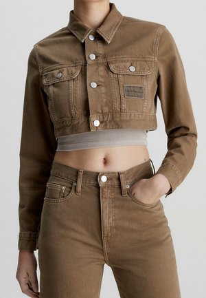 Vrouw draagt een cropped bruine spijkerjas met zilveren knopen en bijpassende hoge taille jeans, bloot middenrif, één hand in de zak.