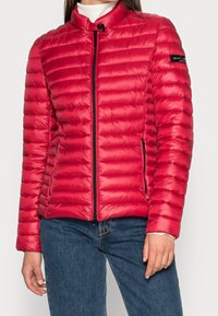 Rød puffjakke med horisontal quiltede design, stående krave, lynlås foran og sidelommer med lynlås. Har en glat, skinnende tekstur.