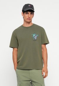 Oliven grøn bomuld t-shirt med korte ærmer, prydet med et grafisk print af en fugl og bogstaver på brystet. Afsluttet med en rund hals.