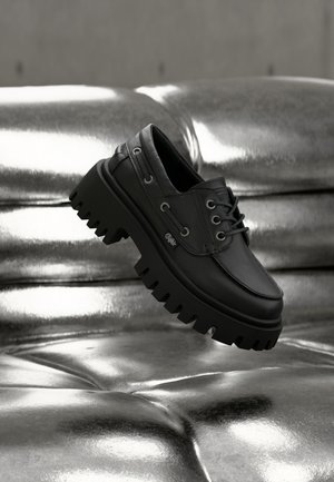 Μαύρο χοντρό πλατφόρμα δετό παπούτσι που αιωρείται πάνω από μια λαμπερή μεταλλική ασημί δερμάτινη επιφάνεια με ραφές τύπου τουφ.