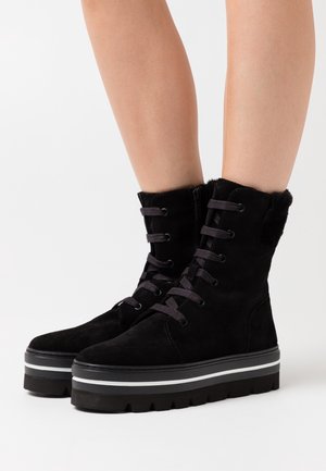 Stivaletti alla caviglia in suede neri con suola spessa nera, strisce bianche, allacciatura frontale e fodera pelosa nella parte superiore. Presentano una cerniera laterale per una facile vestibilità.