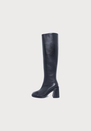 Botte noire en cuir montant jusqu’au genou, à bout arrondi et à talon bloc épais, sur un fond clair uni.