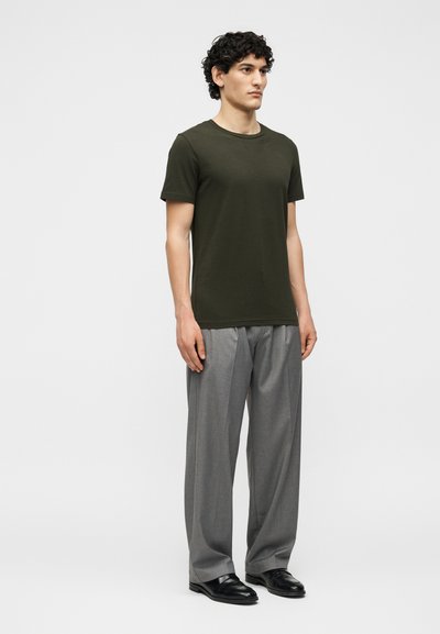 Άνδρας με σγουρά μαλλιά όρθιος, φοράει ελιάς πράσινο κοντομάνικο μπλουζάκι, χαλαρό γκρι παντελόνι και μαύρα παντελονάκια σε μονόχρωμο φόντο.