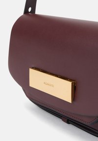 Geantă din piele bordeux cu o textură netedă, având un accent din metal auriu și un închizător rectangular cu numele brandului embosat.