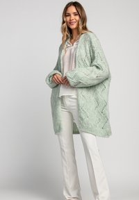 Miętowozielony sweter z dzianiny z otwartym przodem, skomplikowanym wzorem koronkowym i oversize'owymi rękawami, noszony na jasnej bluzce i białych spodniach.