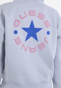 Felpa grigia chiaro con una grande stella blu all'interno di un logo circolare rosa e blu. Il materiale sembra morbido e presenta un design a vestibilità rilassata.