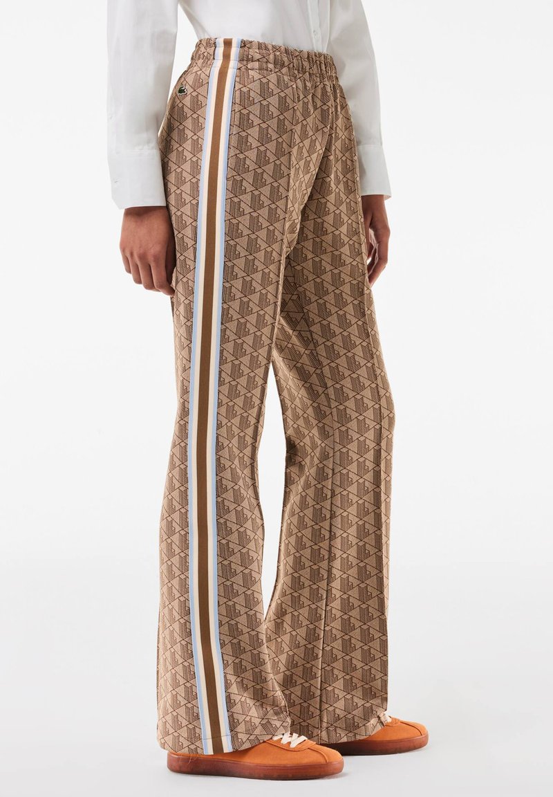 Brown patterned trousers with a geometric design, featuring side stripes in white, blue, and brown. Elastic waistband, flared legs.