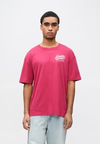 Kortermet, rundhalset t-skjorte i livlig magenta bomull. Har et hvitt logotrykk på brystet og en avslappet passform. Stilfullt kombinert med lys blå jeans.