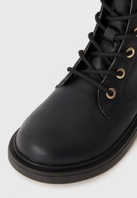 Schwarze Leder-Stiefelette mit runder Zehenpartie, goldfarbenen Metallösen, schwarzen Schnürsenkeln und einer niedrigen, robusten Sohle. Glatte Textur mit minimalen Nähten.