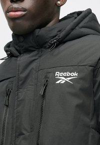 Mustapukuinen Reebok-takki korkealla kauluksella, jossa on vetoketjulliset taskut ja jousto säädettävät hihnat. Logo on selkeästi esillä valkoisena.