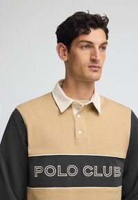Beżowa i czarna długa koszula polo z kremowym kołnierzykiem, z haftem "POLO CLUB" na białym tle z przodu. Materiał bawełniany.
