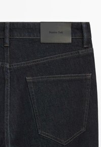 Dark denim jeans with a black leather label, featuring back pockets and gold stitching. Classic cut with a smooth, textured finish.