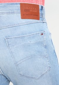 Vaaleansiniset farkut, joissa on takatasku, jossa on hillitty punavalkoinen korostus ja ruskea nahkalappu, jossa lukee "HILFIGER DENIM".