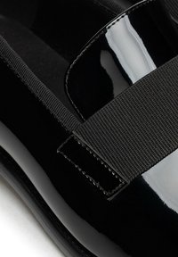 Schwarzer Lacklederschuh mit einem breiten elastischen Riemen am Spann, der eine glatte, glänzende Oberfläche und ein abgerundetes Zehendesign aufweist.