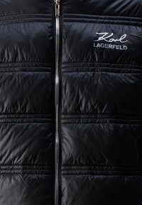 Sort pufferjakke med quiltet tekstur og sølvlynlås. Har "Karl Lagerfeld" logo i hvid på den øverste venstre side.