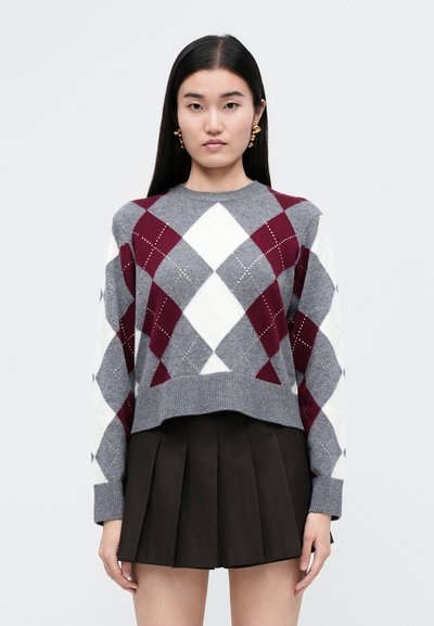 Maglione grigio corto con un motivo a diamante in rosso scuro e bianco, con polsini e orlo a costine. Abbinato a una gonna plissè scura.