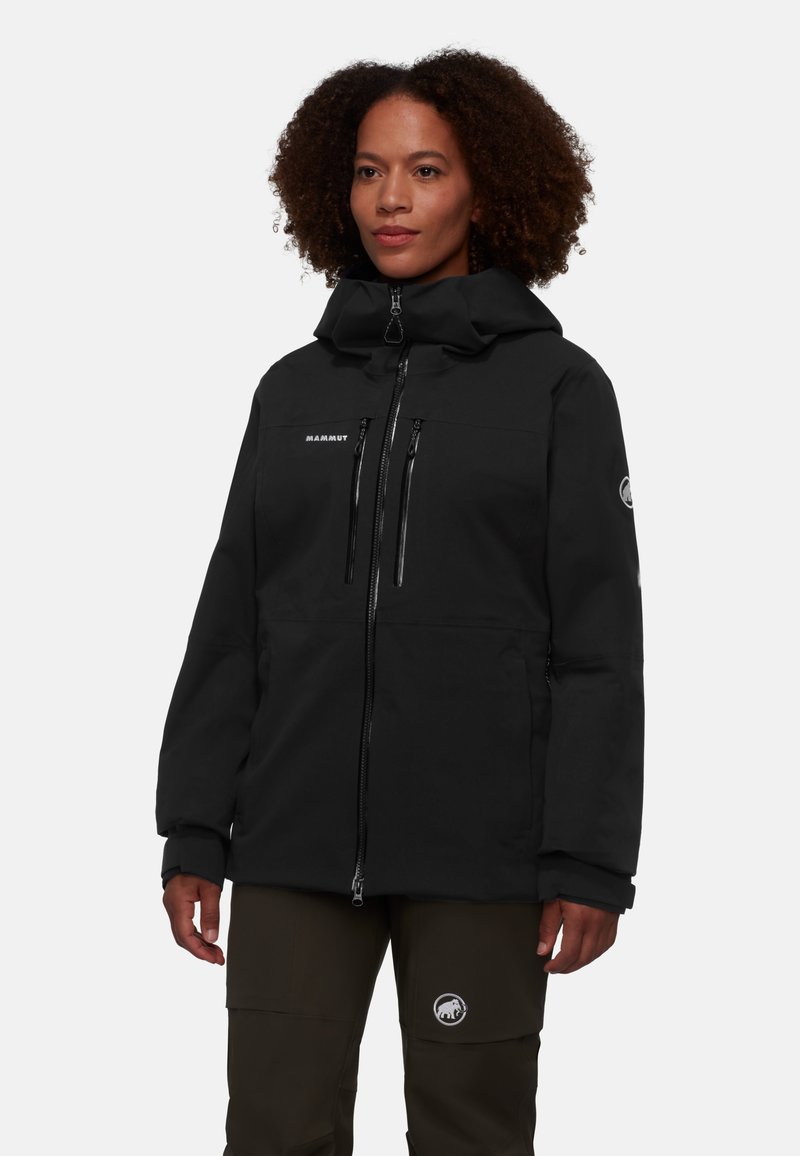 Svart softshelljacka med hög krage, dubbla dragkedjefickor fram och elastiska ärmslut. Bärs med gröna byxor och visar upp en logotyp.