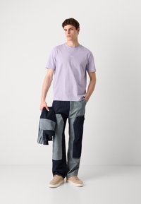 Hellblaues T-Shirt mit zentriertem Logo, kombiniert mit Jeans aus Patchwork in verschiedenen Blautönen. Beigefarbene Sneakers, während sie eine dunkle Jacke hält.