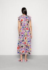 Vestido maxi floral com mangas curtas e volantes, apresentando um padrão de margaridas multicoloridas sobre um fundo roxo. Usado com mules de salto rosas.