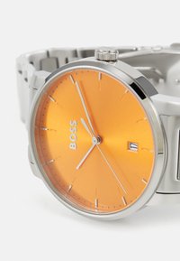 Reloj redondo de acero inoxidable plateado con una esfera metálica naranja, marcadores de hora plateados y una pantalla de fecha a las 3 en punto.