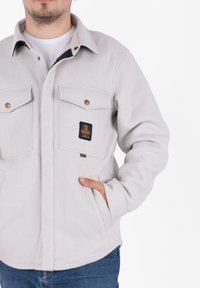 Giacca in pile grigia chiara con colletto, due tasche sul petto e un'etichetta con il marchio. Presenta una chiusura con bottoni e tasche laterali per le mani.