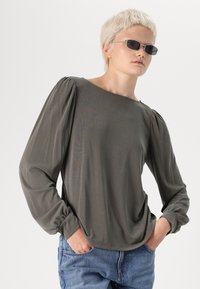 Sötétzöld, hosszú ujjú blúz puffos vállakkal, kerek nyakkivágással és ráncolt mandzsettával, puha, könnyű anyagból. Kék farmerral párosítva.