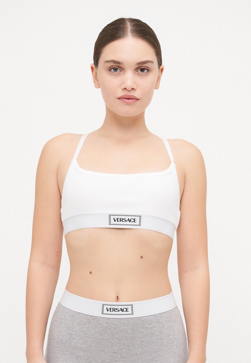 Balta īsā bralette ar plānām siksnām. Iezīmēta ar plašu elastīgu joslu ar melnu "VERSACE" logo. Gluda, elastīga auduma.