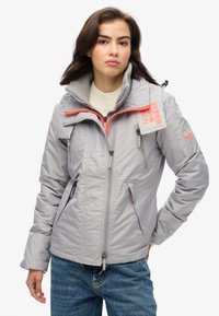 Grå isoleret jakke med orange detaljer, lynlåslommer, høj krave og en tekstureret overflade, parret med blå denim jeans.