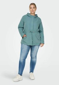 Žalio mėlyno spalvos su kapuce striukė su raišteliu juosmeniui, dviem priekinėmis kišenėmis ir glotnia tekstūra. Dėvima su nusidėvėjusiais mėlynais džinsais ir baltais sportbačiais.