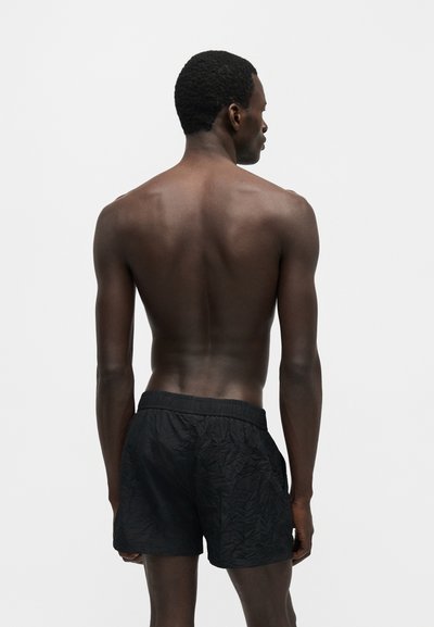 Άνδρας στέκεται πλάτη, χωρίς μπλούζα, φοράει μαύρα υφασμάτινα σορτς μπροστά σε απλό ανοιχτό φόντο.