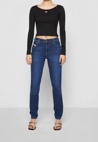 Top corto nero a maniche lunghe con scollatura rotonda e logo, abbinato a jeans attillati in denim blu scuro e sandali neri piatti.