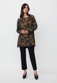 Túnica de manga larga en patrones negro y mostaza con un escote redondo y detalles de botones, combinada con pantalones negros ajustados y zapatos negros.