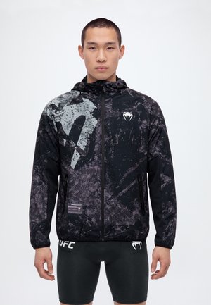 Un hombre posa con una chaqueta con capucha y cremallera de UFC, negra y gris, y unos pantalones cortos negros a juego, sobre un fondo blanco.