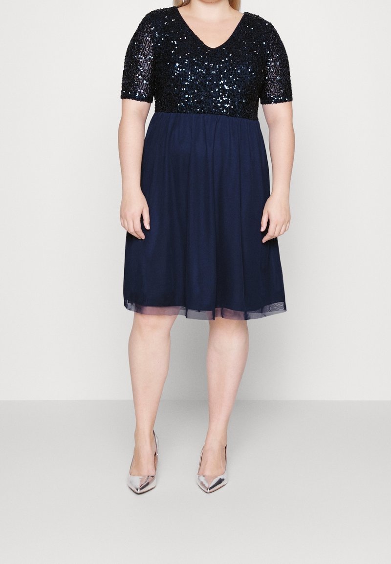 Kobieta ubrana w granatową sukienkę za kolano z cekinowym czarnym topem i krótkimi rękawami, do tego srebrne szpilki z ostrym noskiem.