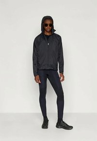 Veste coupe-vent noire avec capuche, associée à un legging noir et des chaussures de sport. Des lunettes de soleil complètent la tenue. Texture lisse avec un design ajusté.