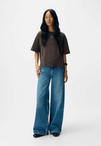 Bruine t-shirt met een blauw logo, gecombineerd met wijde blauwe jeans en zwarte schoenen. Lange donkere haren, eenvoudige uitstraling, felle kleuren. Witte achtergrond.