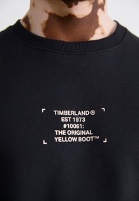 Gros plan d'un t-shirt Timberland noir avec un texte blanc affichant « TIMBERLAND EST 1973 #10061 : THE ORIGINAL YELLOW BOOT™ » sur la poitrine.