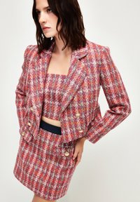 Tweed set bestaande uit een cropped blazer en een getailleerde rok. Voornamelijk roze met multiculturele accenten, metalen knopen en gestructureerde stof.