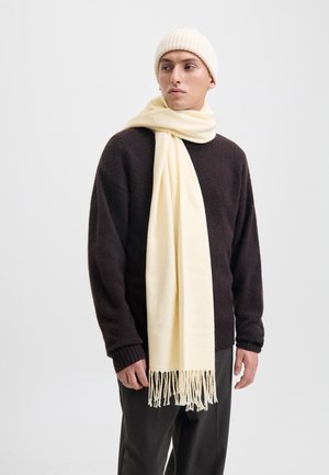 Écharpe crème avec franges drapée autour du cou, associée à un pull texturé marron foncé et un bonnet côtelé de couleur claire.