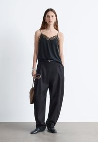Schwarzes Satin-Camisole mit Spitze, kombiniert mit lockeren schwarzen Hosen. Die Accessoires bestehen aus einer kleinen braunen Tasche und silbernen Armbändern.