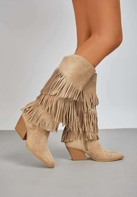 Bottines en daim beige avec des accents frangés et un bout pointu. Comprend un talon empilé et une surface texturée.